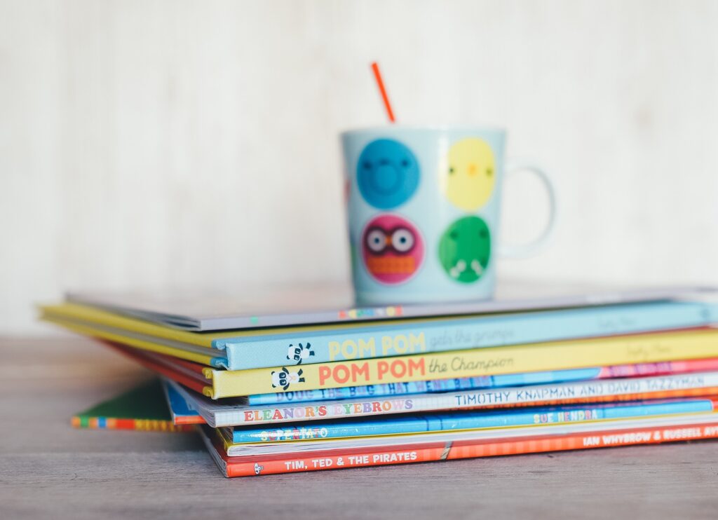 Barevné dětské knihy naskládané na sebe a na nich modrý hrnek se smajlíky.