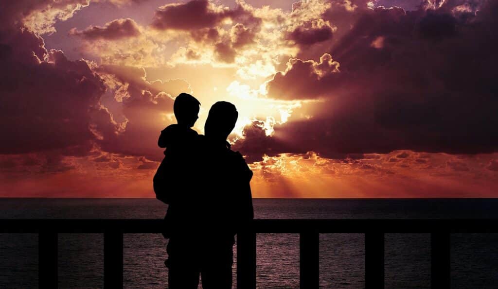 Otec drží v ruce syna a stojí na molu při západu slunce.