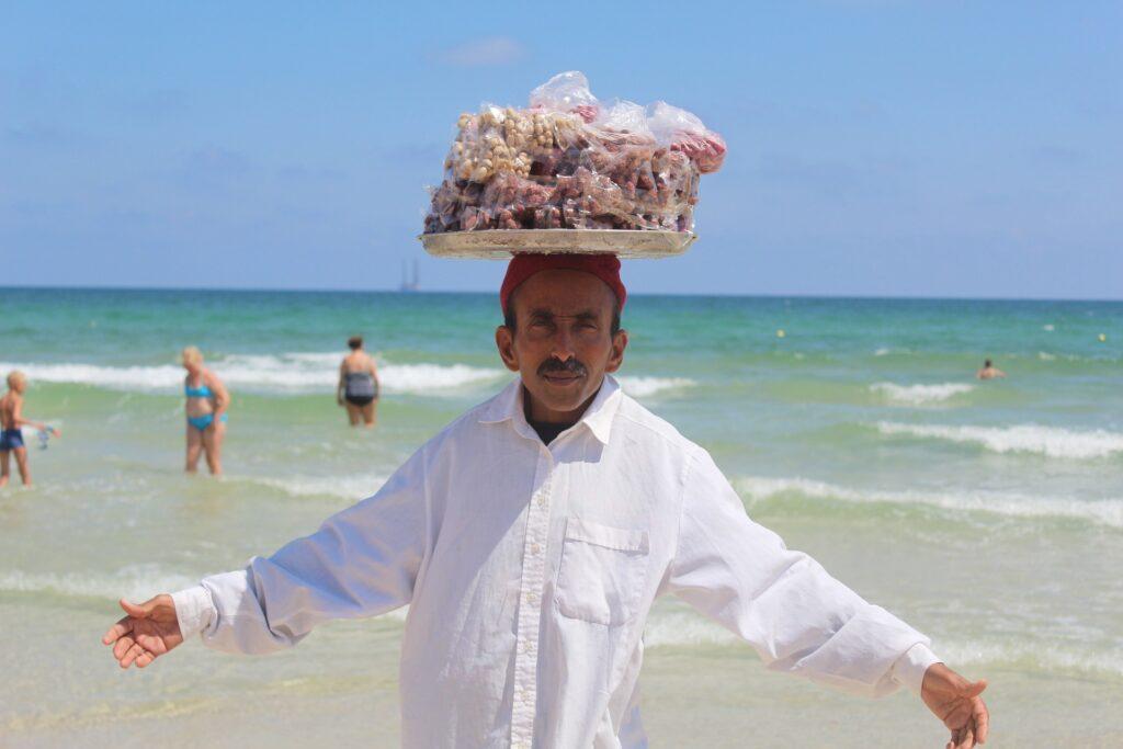 Domorodec má na hlavě tác s jídlem a stojí na pláži.